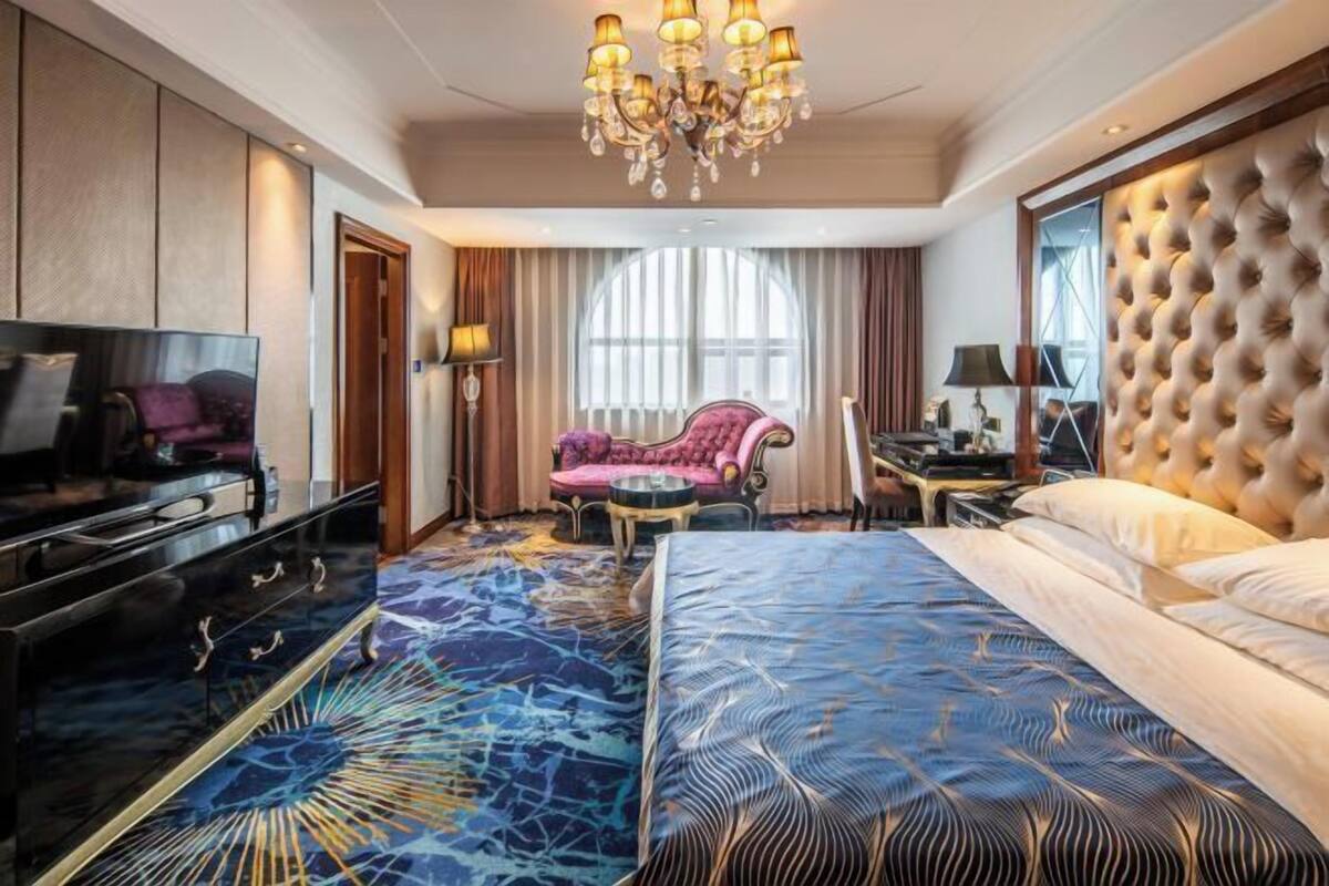 A hotel room with a large bed, a television, a seating area with a chair, and a chandelier.