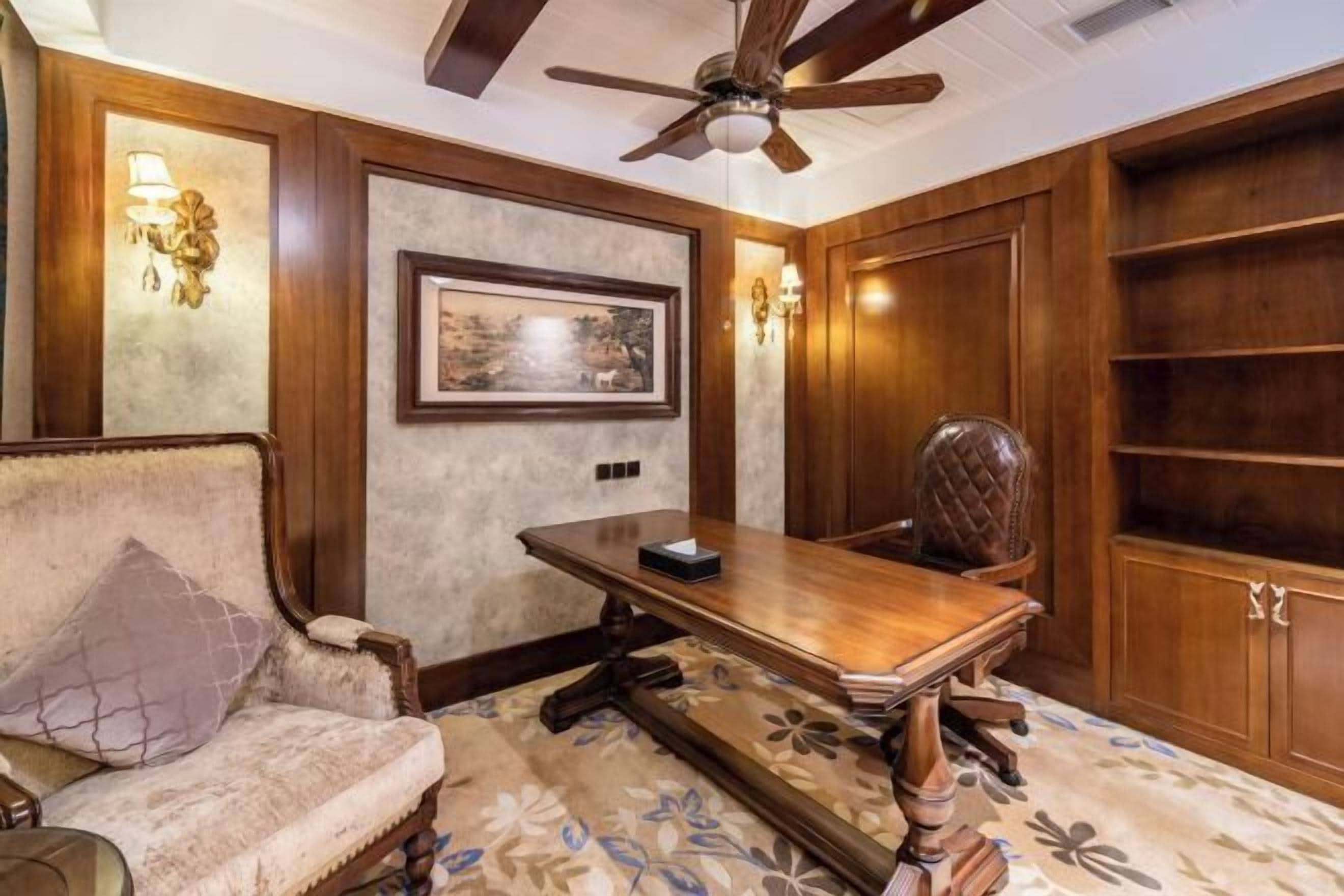 Une pièce avec un bureau en bois, une chaise rembourrée, un ventilateur de plafond et un tableau encadré au mur.