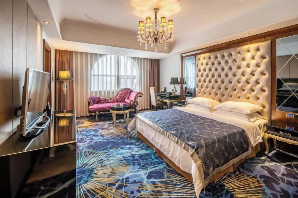 A hotel room with a large bed, a TV, a pink armchair, a desk, and a chandelier.