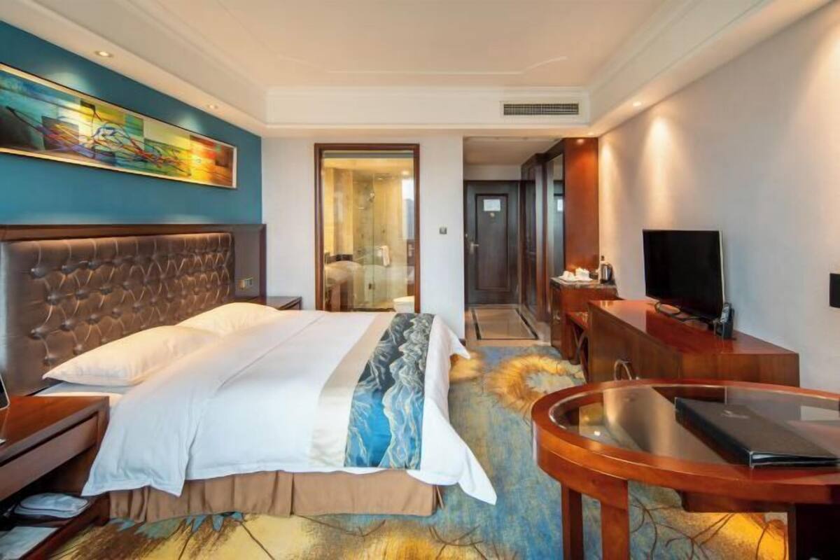 A hotel room with a large bed, a desk with a TV, a bathroom, and a painting on the wall.