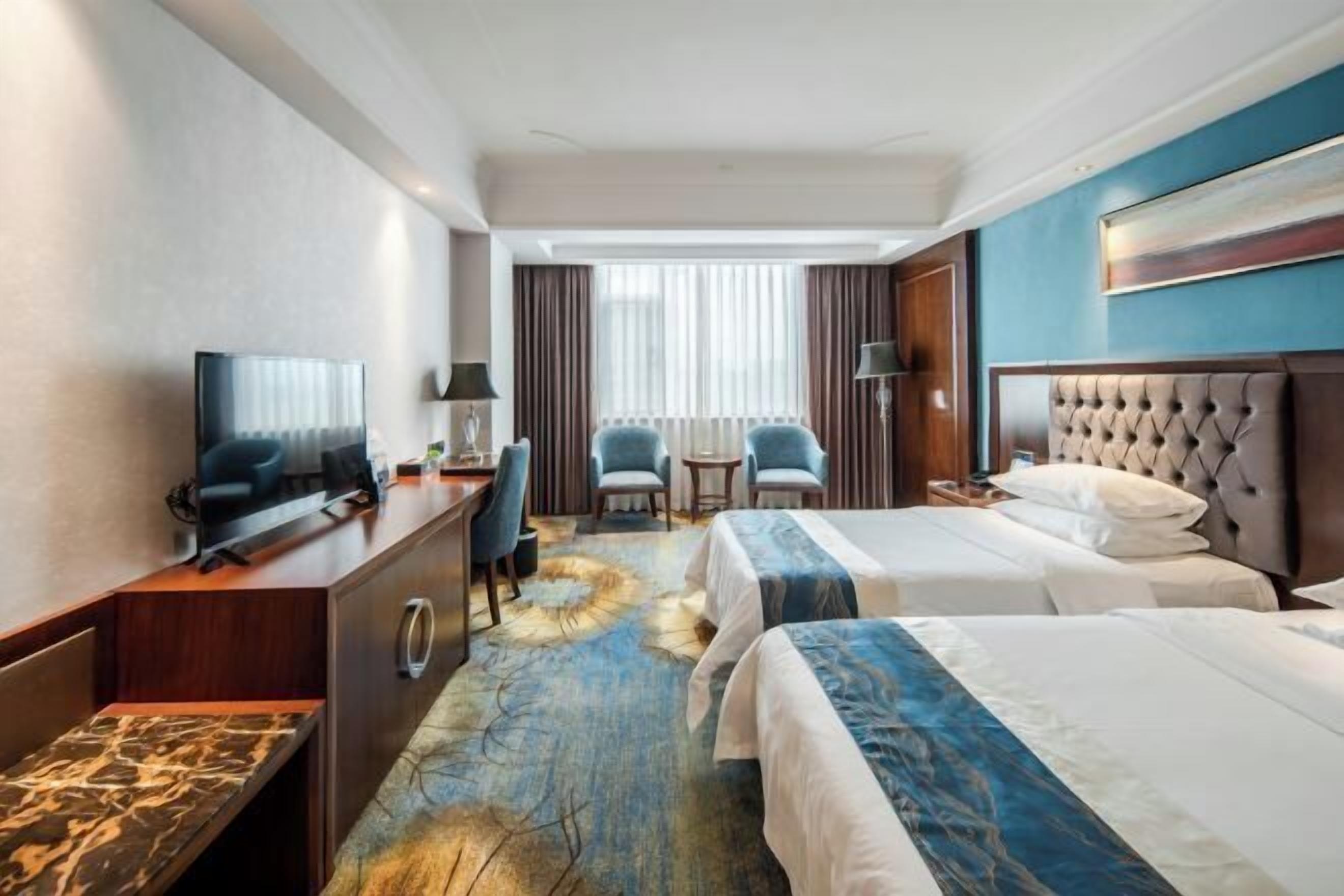 A hotel room with two beds, a desk with a television, a chair, and a view of the outdoors through large windows.