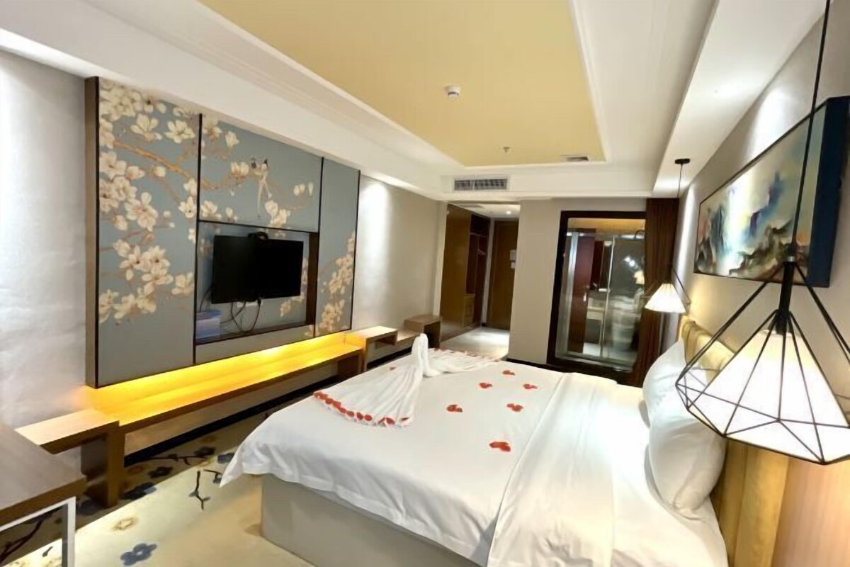 A hotel room with a bed, a TV mounted on the wall, a desk, and a lamp.