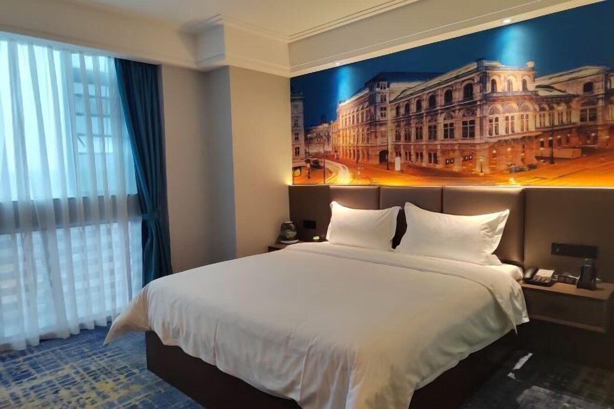 A hotel room with a large bed, a nightstand, a lamp, and a painting of a building on the wall.