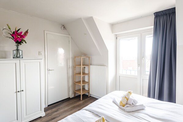 A bedroom with a bed, a wardrobe, a radiator, a wooden shelf unit, and a window with curtains.