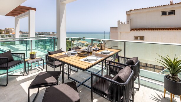 Ein Balkon mit Holztisch, gedeckten Plätzen, schwarzen Metallstühlen, einer Glastrennwand und Blick aufs Meer.