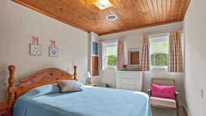 Ein Schlafzimmer mit einem Bett mit Holzkopfteil, einem Nachttisch mit Lampe, einem Sessel mit rosa Kissen und einem Fenster mit Vorhängen.