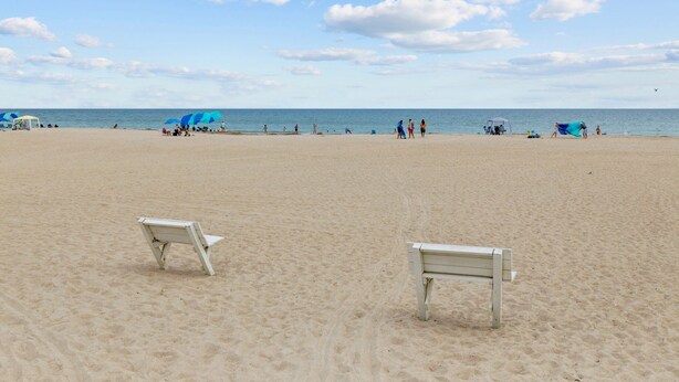 Ein Strand mit zwei leeren weißen Bänken, wolkenlosem Himmel und dem Meer im Hintergrund.