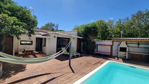 Un patio trasero con terraza de madera, una hamaca, una piscina y una zona de barbacoa.