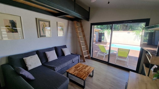 Una sala moderna con sofá, una mesa de centro de madera y un amplio ventanal corredero que da a un espacio exterior con piscina y mobiliario de jardín.