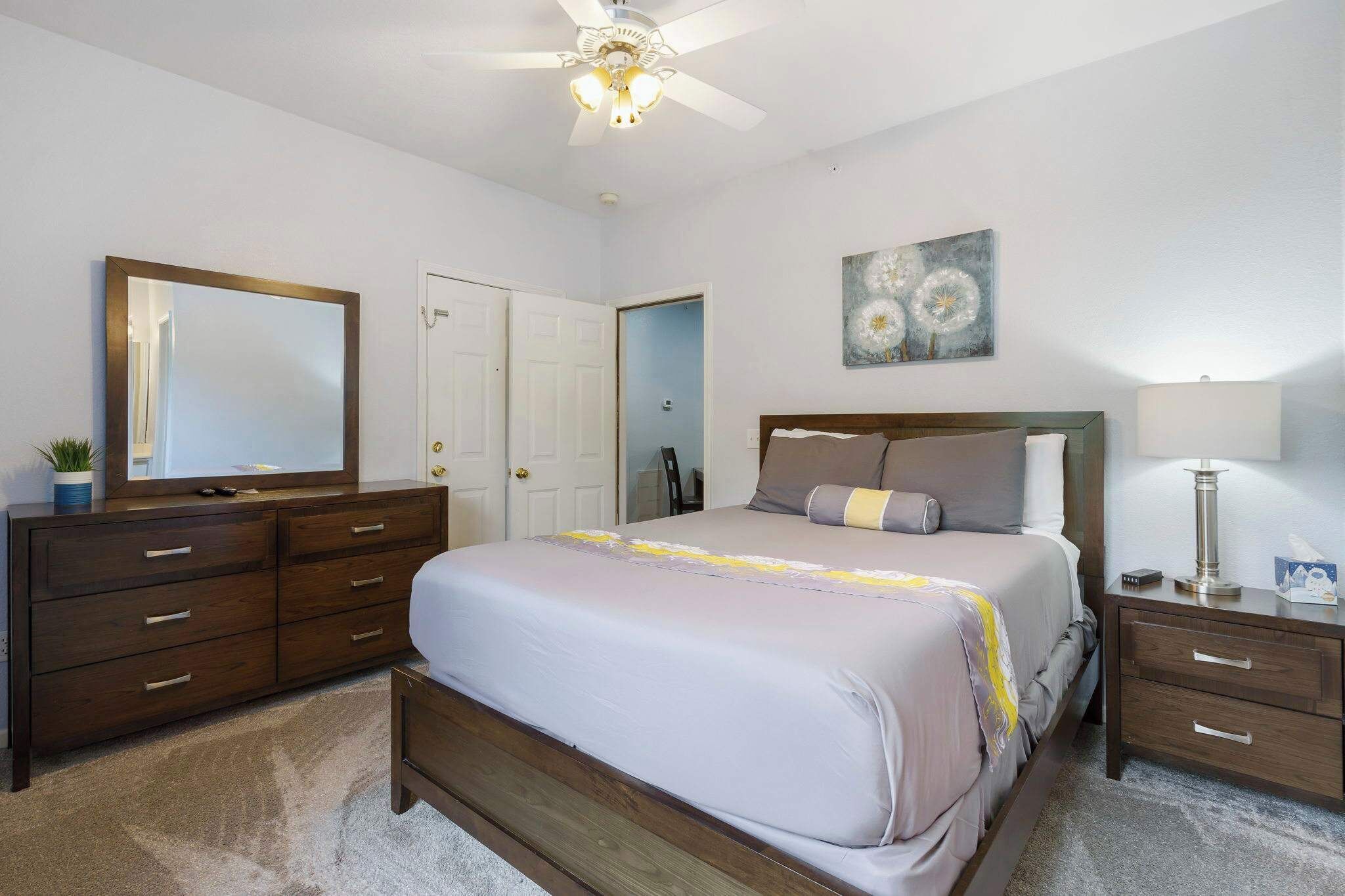 A bedroom with a bed, dresser, nightstand, ceiling fan, and a painting on the wall.