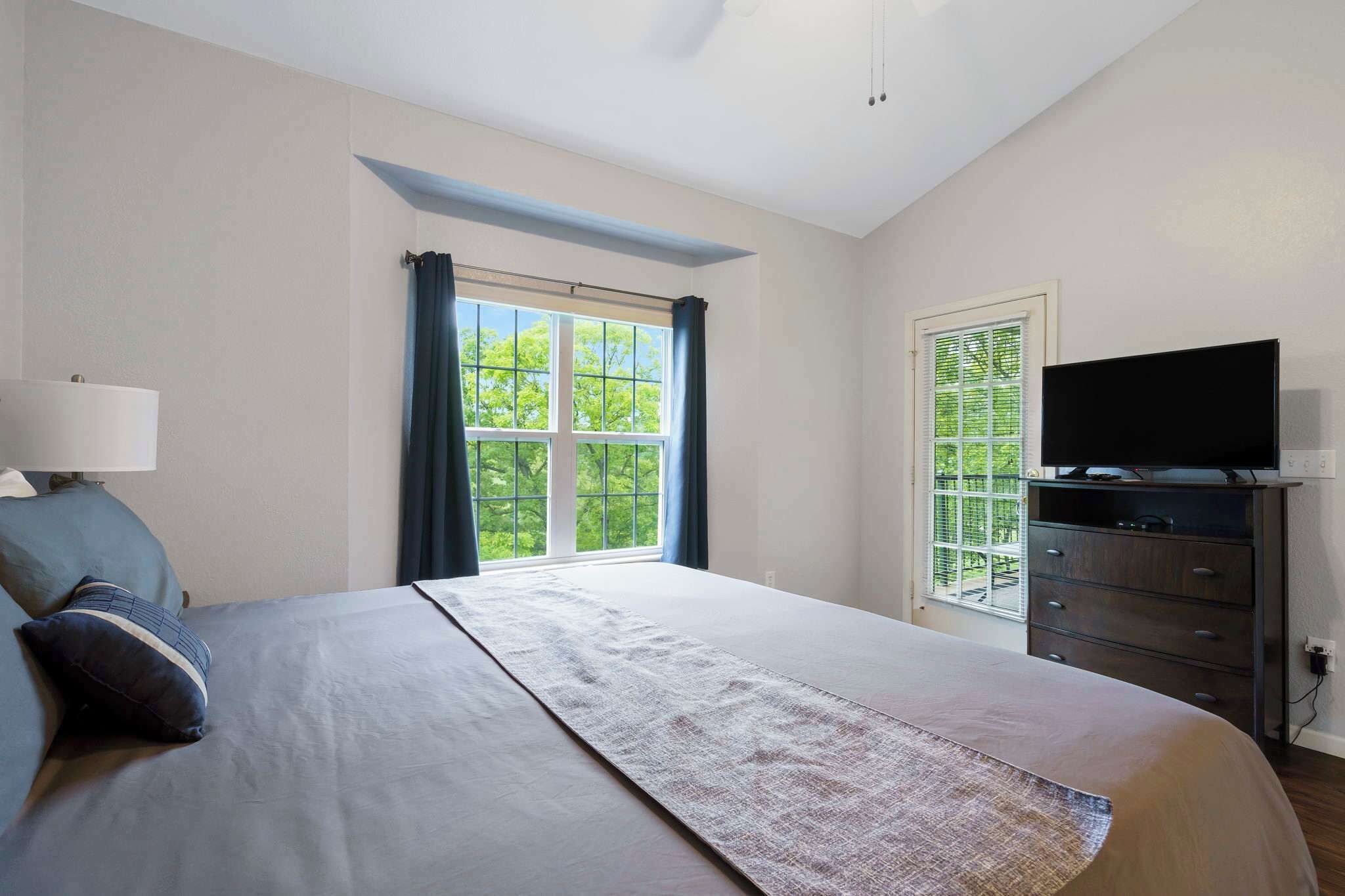 Un dormitorio con cama, televisor en una cómoda, ventana con cortinas y vista a vegetación.