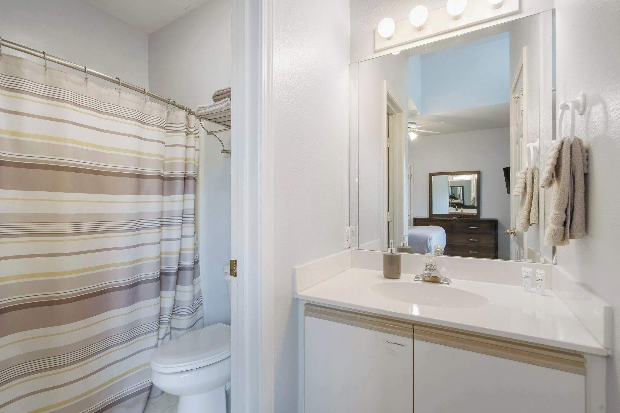 A bathroom with a striped shower curtain, a white sink, a mirror with lights, and a towel rack.