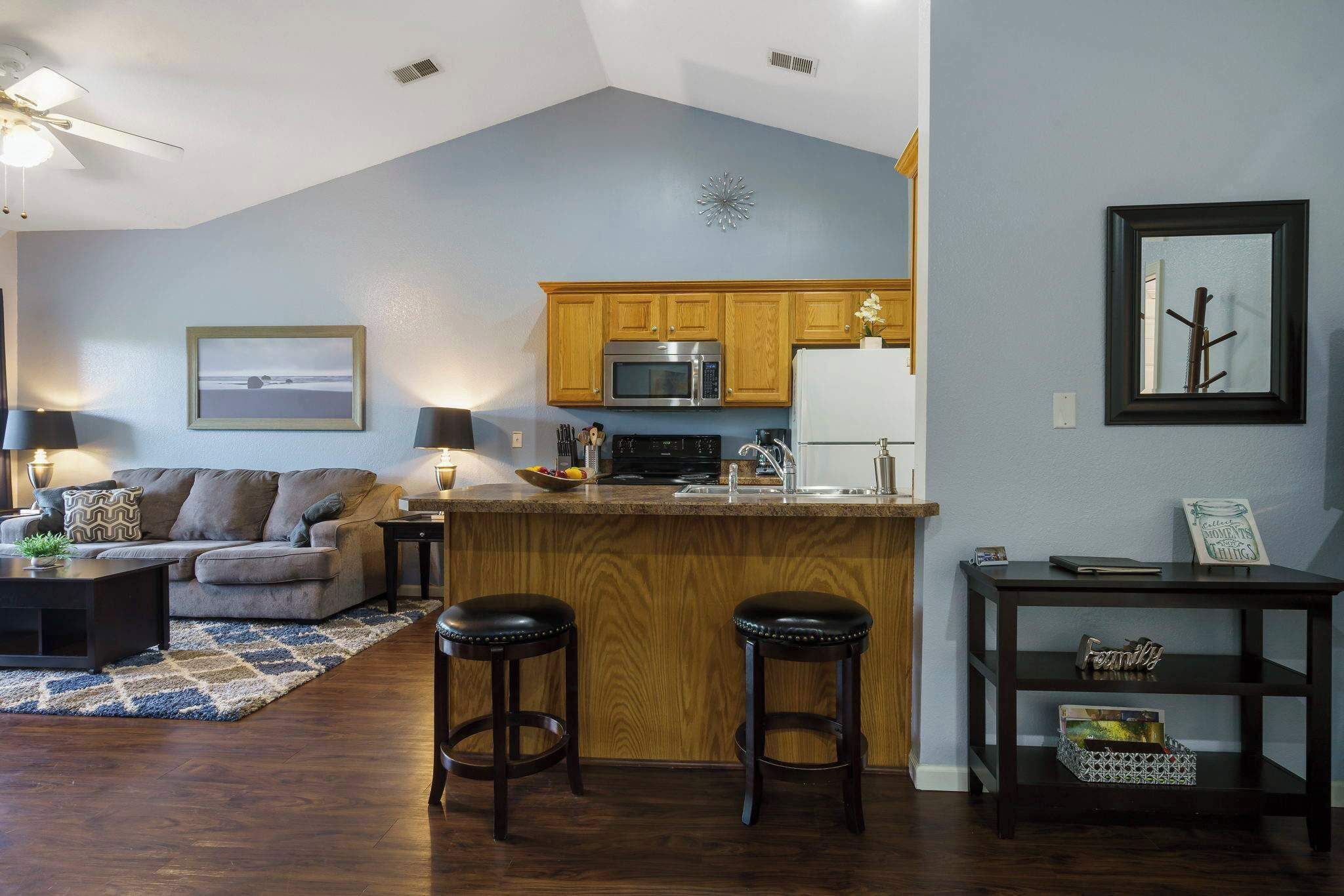 A modern kitchen with wooden cabinets, a microwave, and a refrigerator. There is a bar area with stools and a small table with a decorative item.