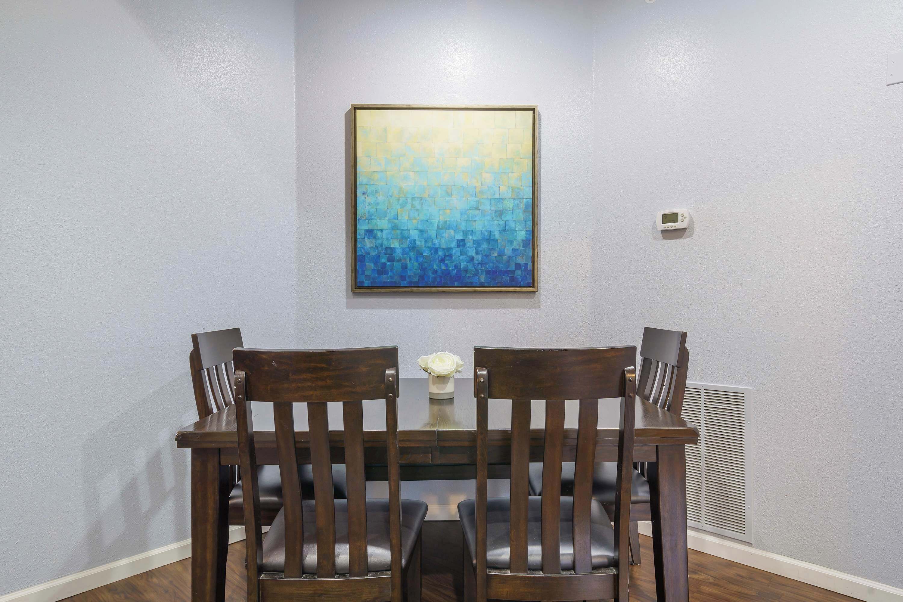 A dining area with a table and chairs, a painting on the wall, and a thermostat on the wall.