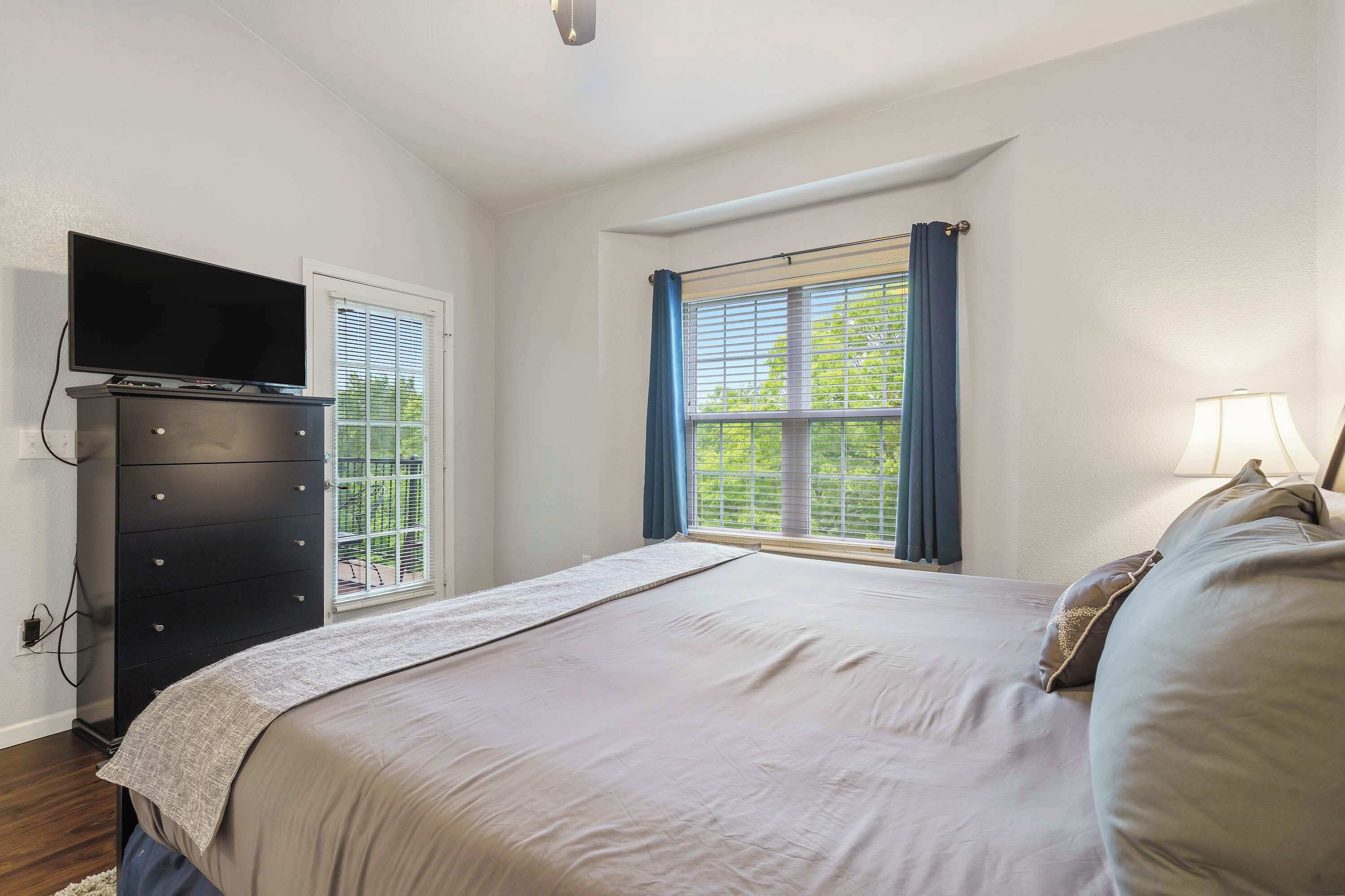 A bedroom with a bed, a dresser, a television on the dresser, a window with curtains, and a lamp on the bedside table.