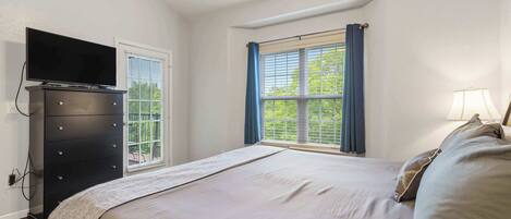 A bedroom with a bed, a dresser, a television on the dresser, a window with curtains, and a lamp on the bedside table.