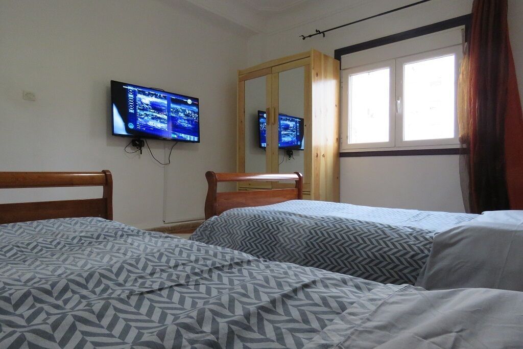 Une chambre avec deux lits, un téléviseur fixé au mur, un miroir et une fenêtre avec des rideaux.
