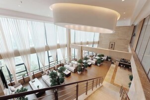 Un amplio vestíbulo de hotel de varios niveles con diseño moderno, que presenta una zona de estar central, ventanas grandes y una escalera.