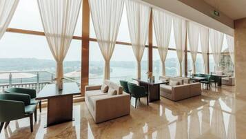 Amplia habitación con vista al mar, que cuenta con una zona de estar con sofá y sillones, una mesa y ventanas grandes con cortinas traslúcidas.