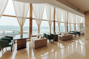 Amplia habitación con vista al mar, que cuenta con una zona de estar con sofá y sillones, una mesa y ventanas grandes con cortinas traslúcidas.