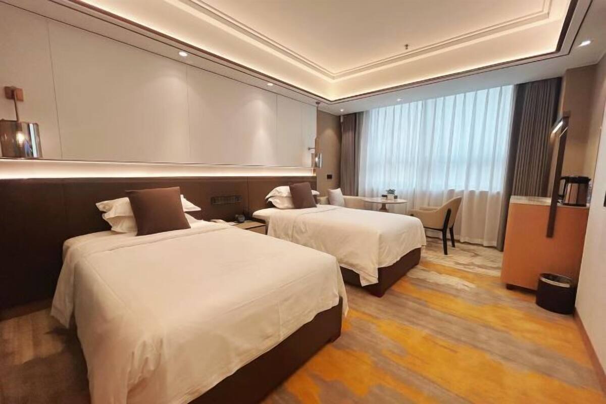Habitación de hotel con dos camas, un escritorio, una silla y un ventanal grande con cortinas.