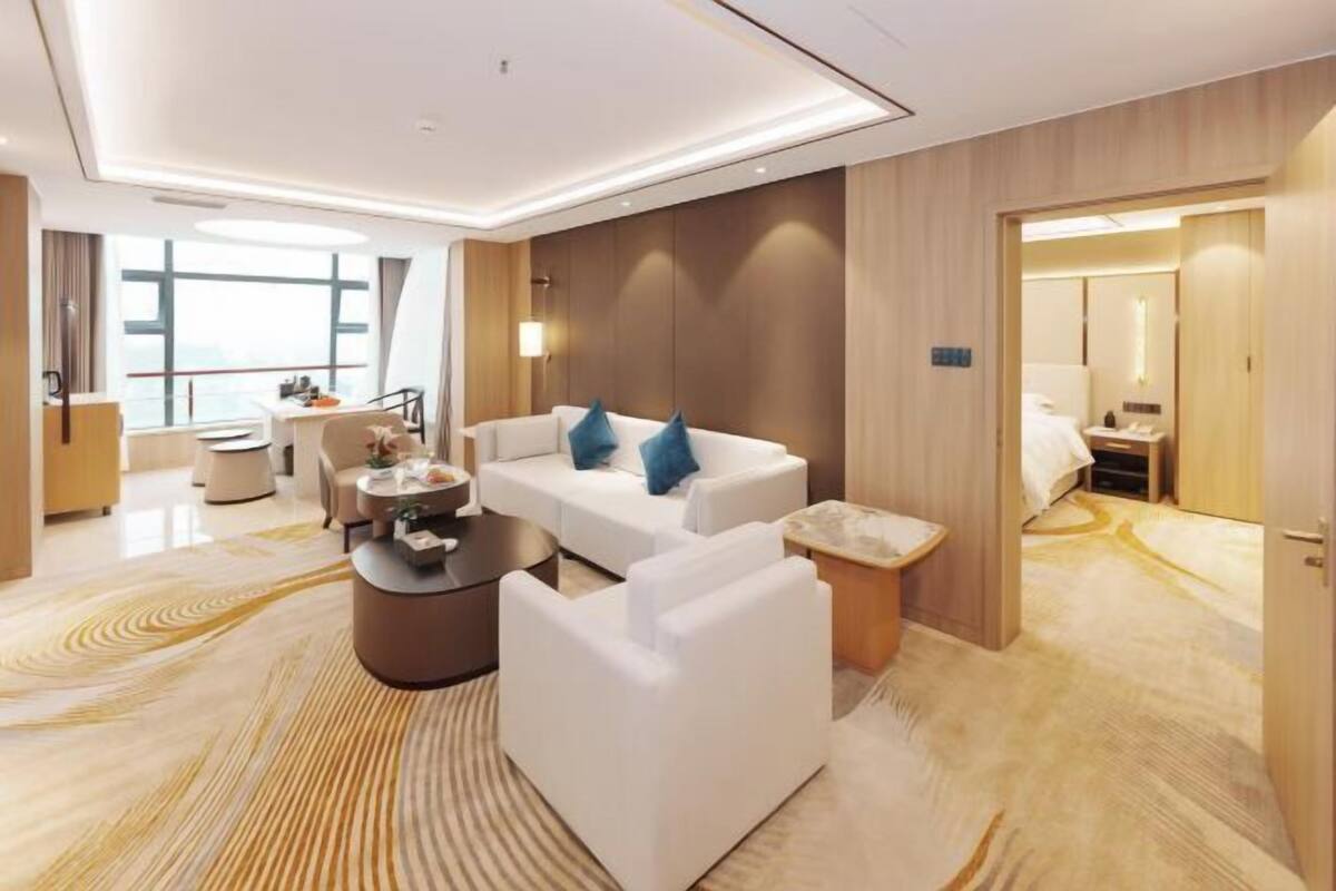Una habitación de hotel moderna con sofá, sillón y una mesita. Se observa una cama al fondo y vistas de la ciudad a través de amplios ventanales.