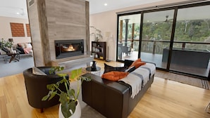 A modern living room with a fireplace, a large window with a view of greenery, and a balcony with outdoor furniture. - Rivers Edge: Experience a truly unique and serene holiday (Toorloo Arm)