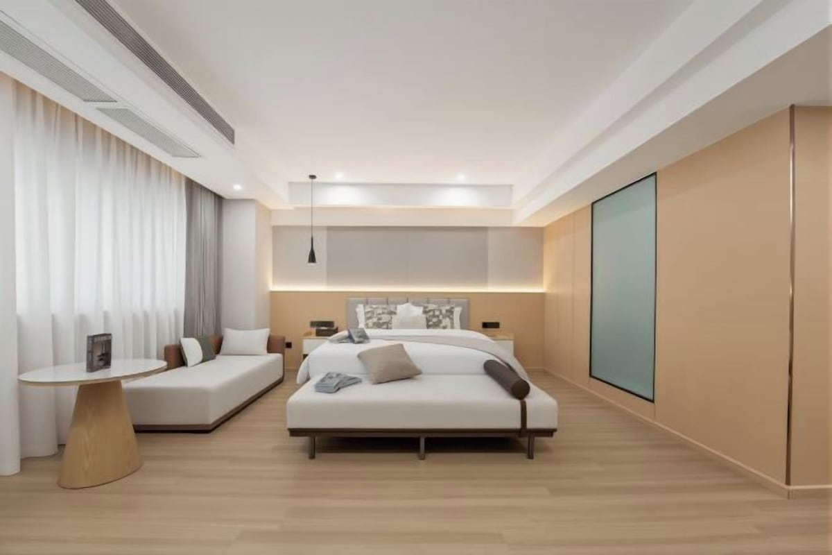 A modern hotel room with a large bed, a sofa, a small table, and a large window.