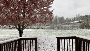 Una terraza cubierta de nieve con baranda de madera, un árbol con hojas rojas y un campo de césped al fondo.