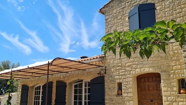 Un bâtiment en pierre avec un toit en tuiles, une porte en bois et un patio couvert aménagé avec du mobilier d’extérieur.