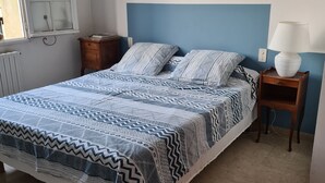 Une chambre à coucher comprenant un lit, une table de chevet, une lampe, une fenêtre avec des rideaux et une estampe botanique encadrée accrochée au mur.