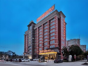 A multi-story hotel with a prominent sign at the top, a ground-level entrance with a lit sign, and a parking lot with several cars.