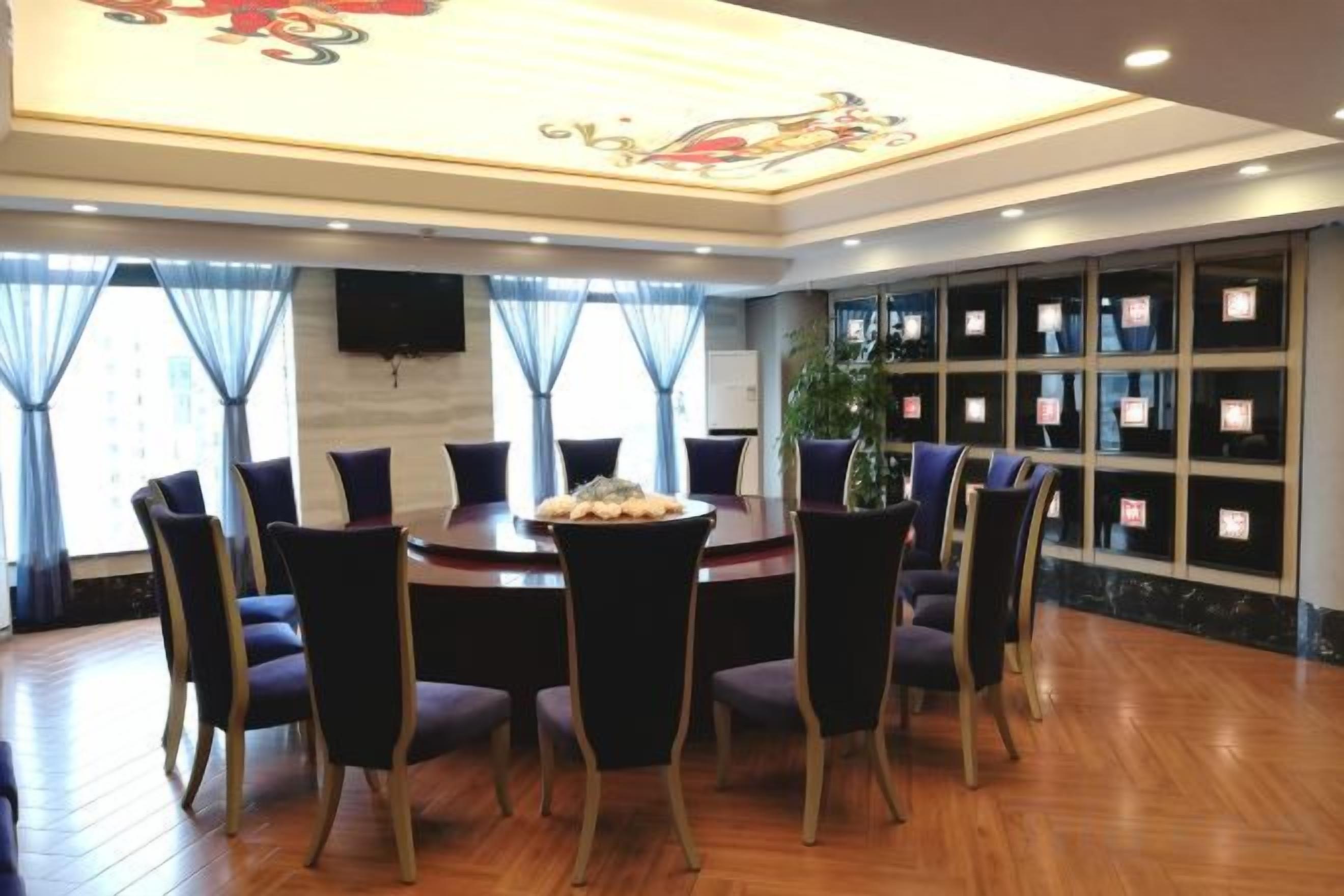 Un salón comedor de hotel con una mesa redonda rodeada de sillas, un ventanal con cortinas y un televisor montado en la pared.