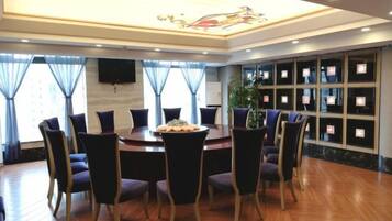 Un salón comedor de hotel con una mesa redonda rodeada de sillas, un ventanal con cortinas y un televisor montado en la pared.