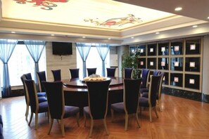 A hotel dining area with a round table surrounded by chairs, a large window with curtains, and a wall-mounted television. - Tiancheng Plaza Hotel (Neijiang)