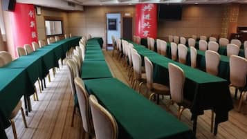 Un salón de conferencias con filas de mesas y sillas, un escenario y un estandarte rojo con texto blanco.