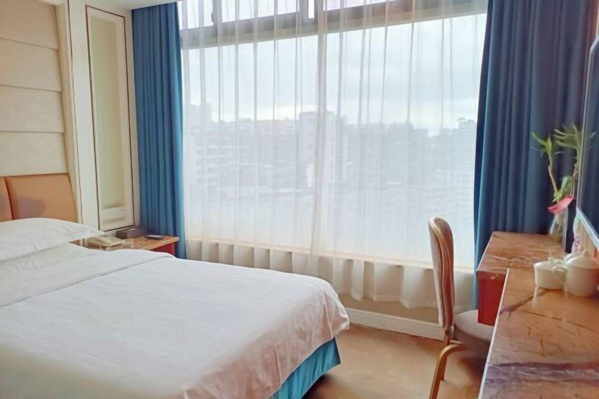 Ein Hotelzimmer mit Bett, Schreibtisch, Stuhl, einem Fenster mit Vorhängen und Blick auf Gebäude.