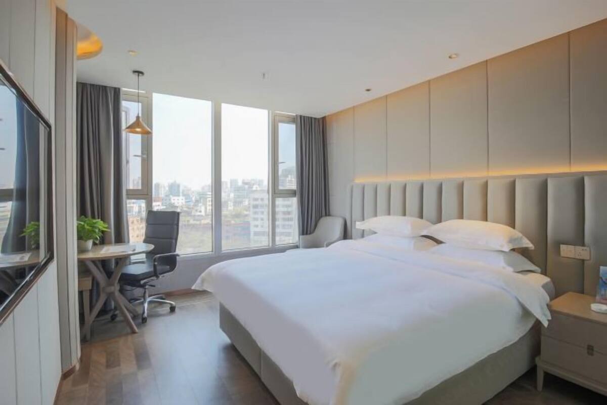 Ein modernes Hotelzimmer mit einem großen Bett, einem Schreibtisch, einem Sessel und einem großen Fenster mit Blick auf die Stadt, sowie ein kleiner Tisch mit einer Pflanze.