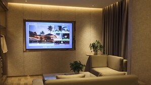 A modern hotel room with a large flat-screen TV mounted on the wall, a comfortable sofa, a small side table with a plant, and a curtain-covered window. - Tiancheng Plaza Hotel (Neijiang)