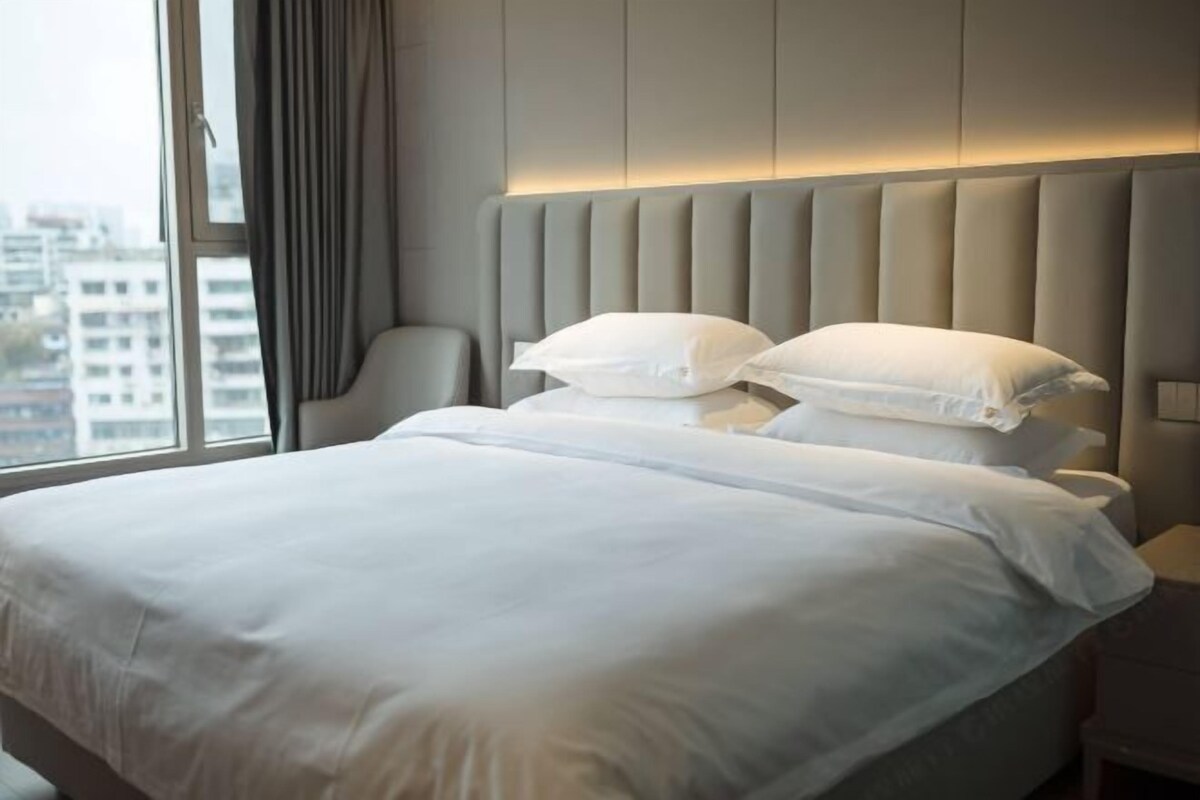 Een hotelkamer met een groot bed, een hoofdbord met verticale panelen, een stoel en een uitzicht op gebouwen door een raam.