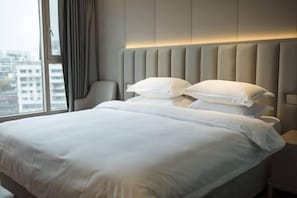 Habitación de hotel con una cama grande, cabecera con paneles verticales, una silla y vistas de edificios a través de una ventana.