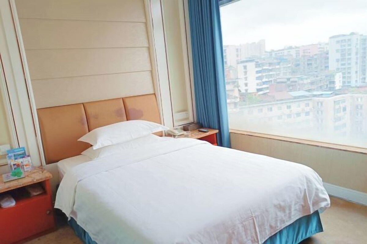 Een hotelkamer met een bed, een nachtkastje, een raam met gordijnen en uitzicht op gebouwen.