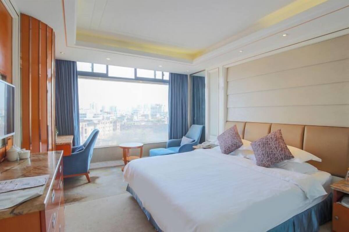 Ein Hotelzimmer mit einem großen Bett, einem Schreibtisch, einem Sessel und Blick auf die Stadt durch das Fenster.
