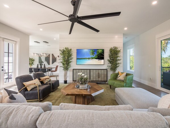 A modern living room with a ceiling fan, a large flat-screen TV, and a dining area with a table and chairs. - North Shore Beach House: Tranquil 4BR Retreat with Pool, Spa, & Walk to Beach (Anna Maria)