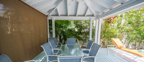A covered outdoor patio with a glass-top table and blue chairs, surrounded by lush greenery and a pool.