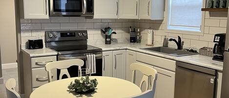 A modern kitchen with white cabinets, stainless steel appliances, a round dining table with chairs, and a window with blinds.