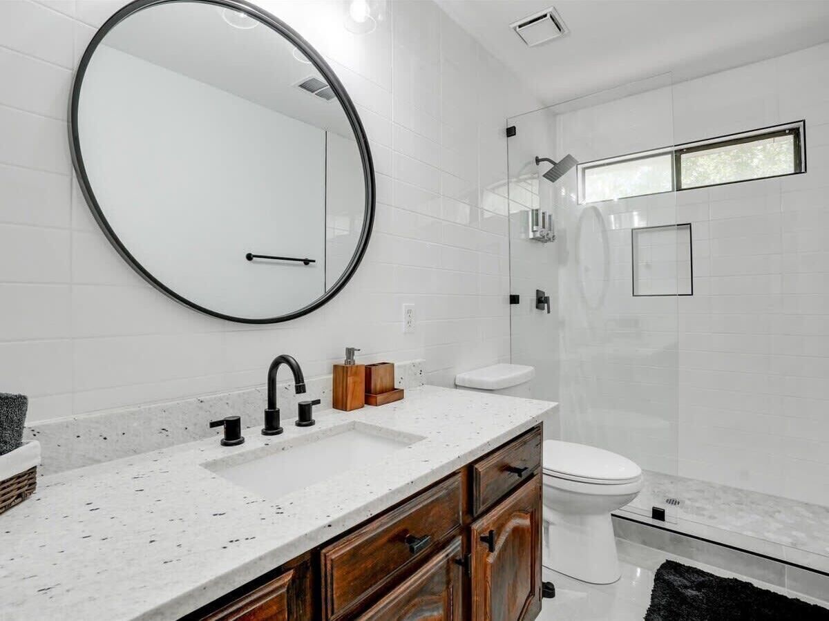 A modern bathroom with a large round mirror, a white sink, wooden cabinets, and a white toilet.