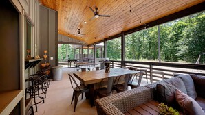 Un área de comedor al aire libre techada con una mesa y sillas de madera, un sofá de mimbre y vistas al bosque.