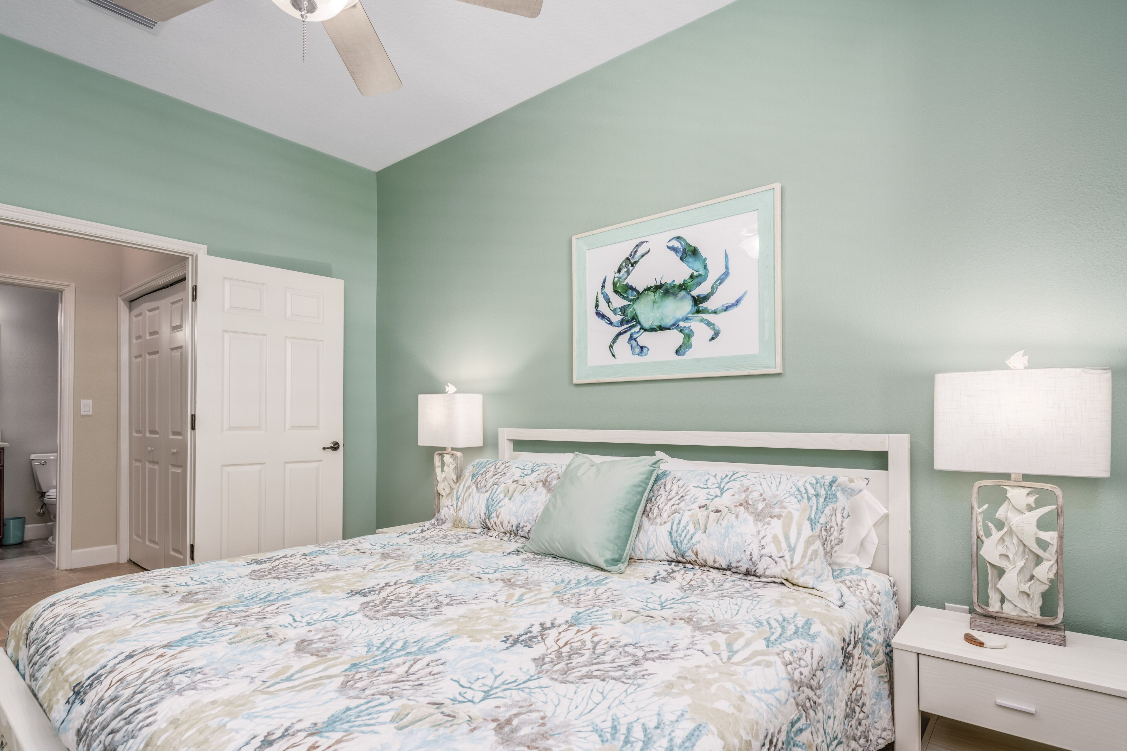 Quarto com uma cama, duas luminárias de cabeceira, uma obra de arte emoldurada de um caranguejo e uma porta que dá para outro cômodo.
