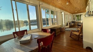 Une pièce spacieuse dotée de grandes fenêtres donnant sur le lac, d’un plancher en bois et d’un coin repas avec une table et des chaises.